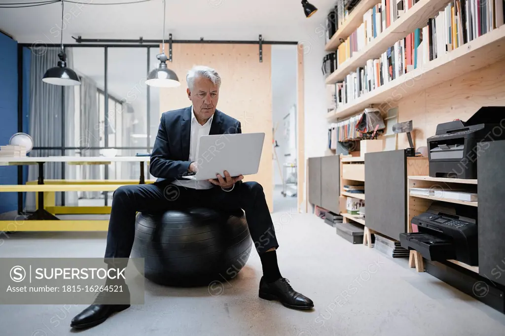 Senior businessman sitting on fitness ball, using laptop