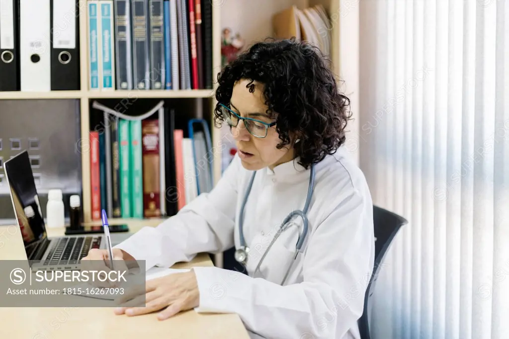 Female doctor writing by laptop while sitting at desk in office