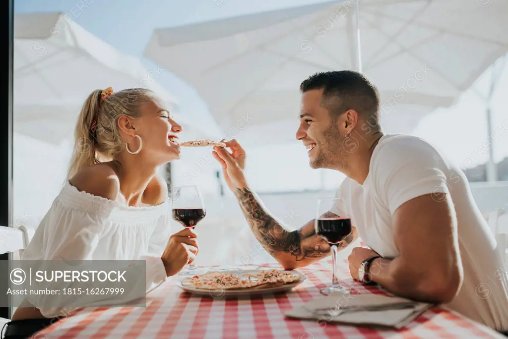 Happy young man feeding pizza to girlfriend while sitting in restaurant