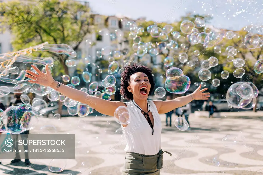 Carefree mid adult woman playing with bubbles while standing in city