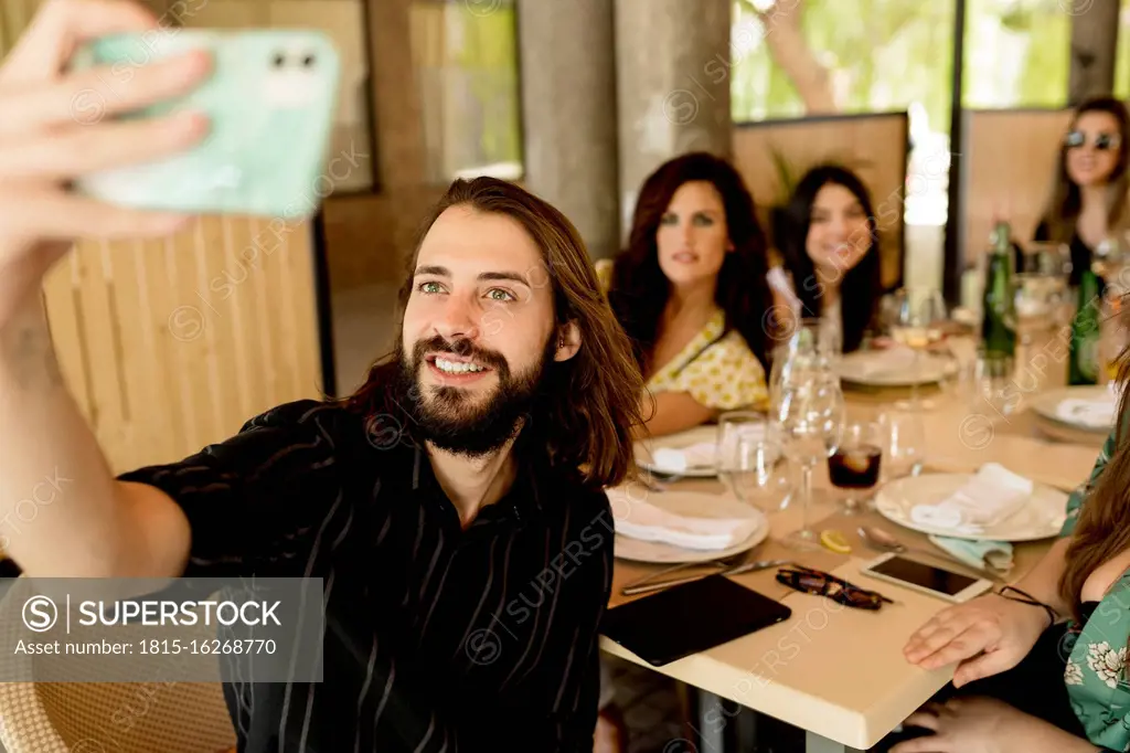 Smiling young man taking selfie with female friends sitting at table in restaurant