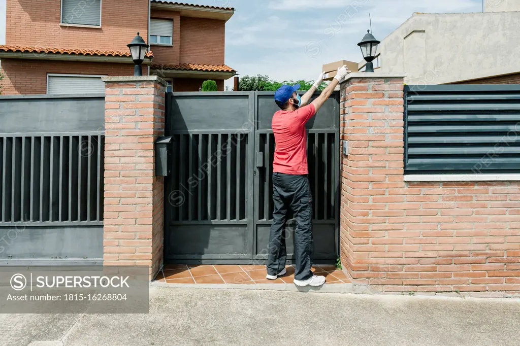 Male postal worker delivering package from over house gate while maintaining social distancing during COVID-19