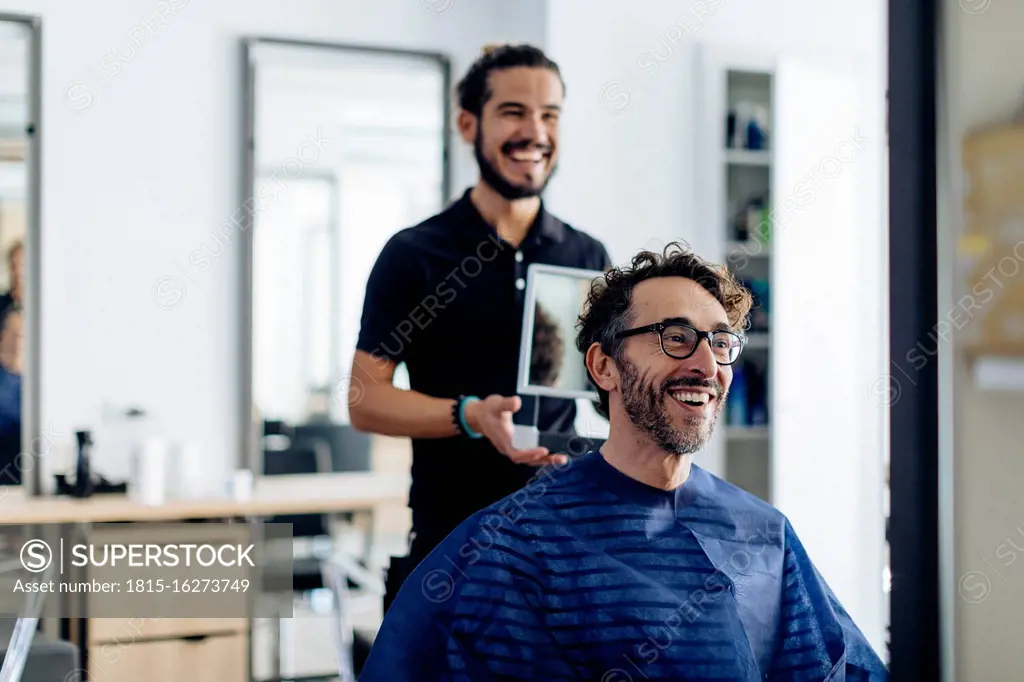 Happy hairdresser showing haircut to client in mirror at salon
