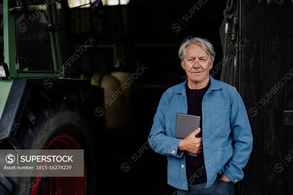 Portrait of a confident senior man holding tablet on a farm with tractor in barn