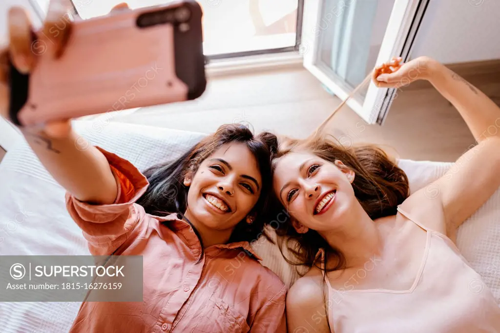 Friends taking selfie with mobile phone while lying on bed at home