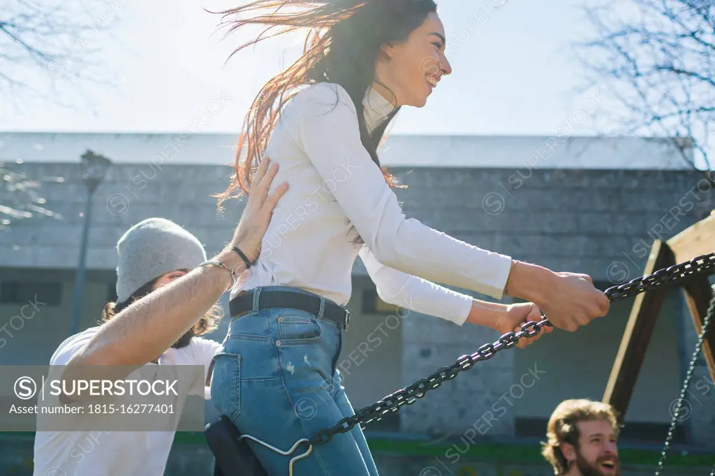 Young man pushing playful friends swinging in park during sunny day