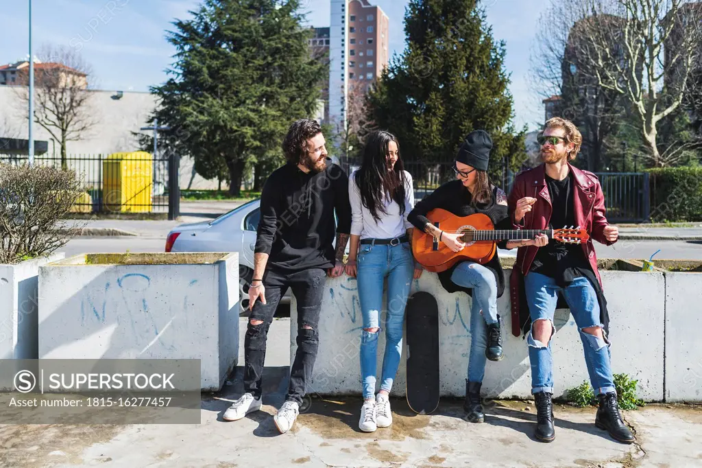 Woman playing guitar while standing with cheerful friends by retaining wall in city