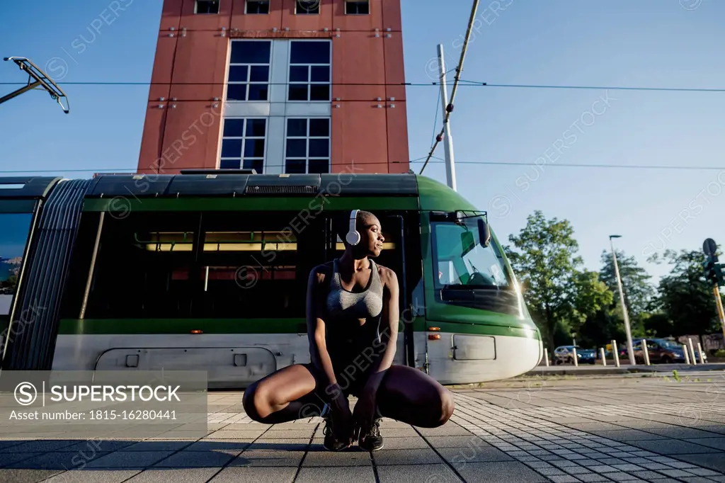 Female athlete listening music through headphones while crouching on street in city