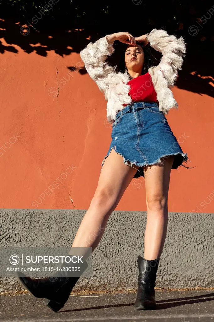 Stylish young woman standing on road against orange wall in city during sunny day