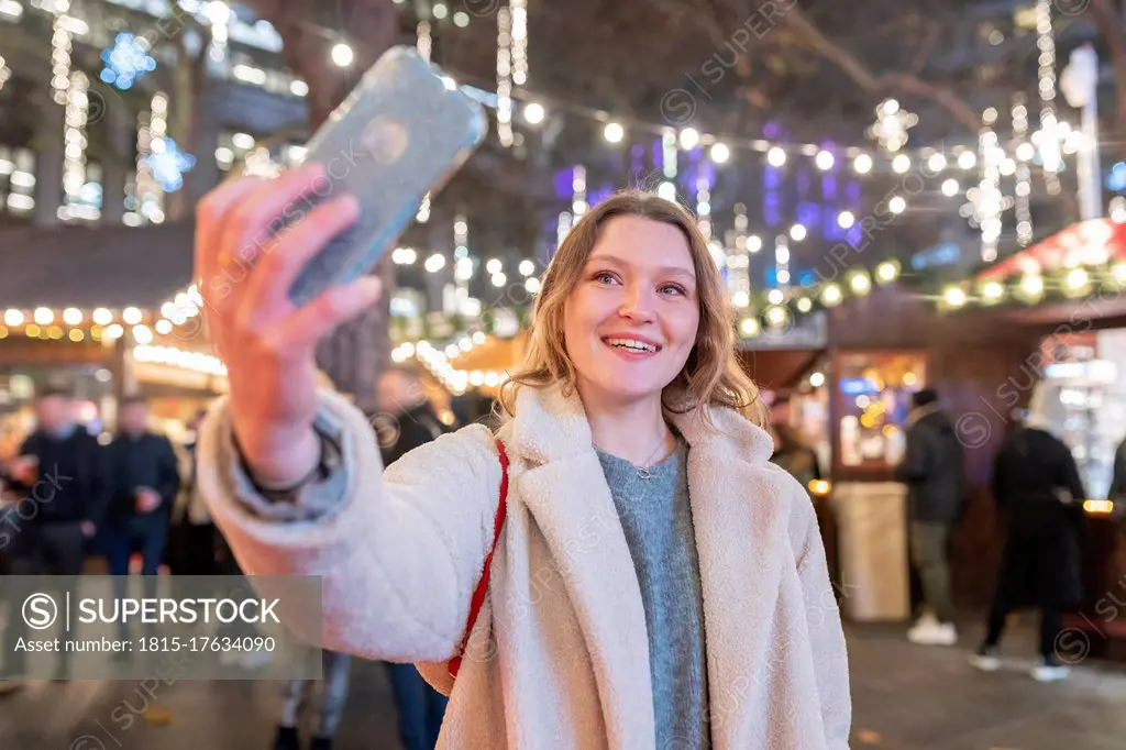 Young woman taking selfie while standing in illuminated Christmas market at night