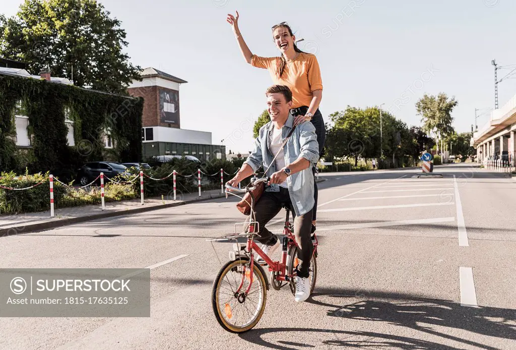 Cheerful young woman standing behind boyfriend riding bicycle on street