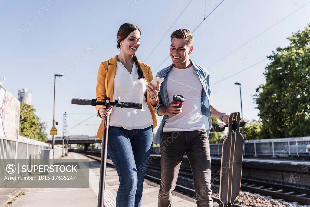 Smiling woman showing smartphone to male friend while walking at railroad station