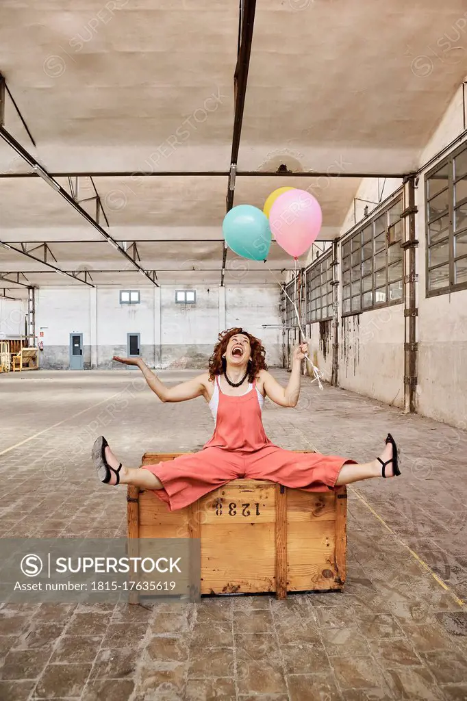 Cheerful woman playing with colorful helium balloons while sitting on wooden box