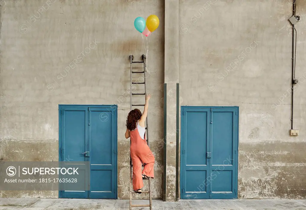 Woman climbing ladder leaning on wall while reaching for colorful helium balloons