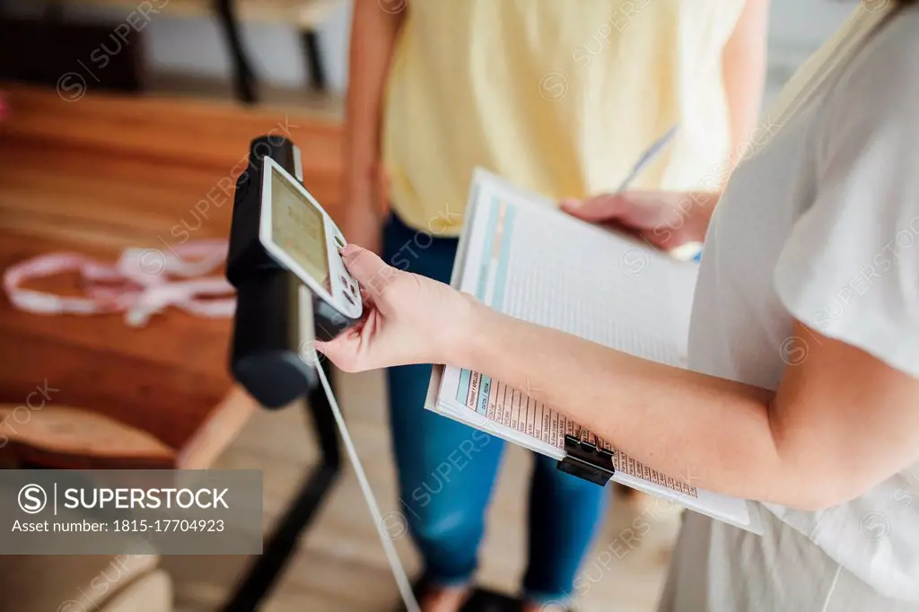 Young woman making diet plan by measuring friend's weight at home