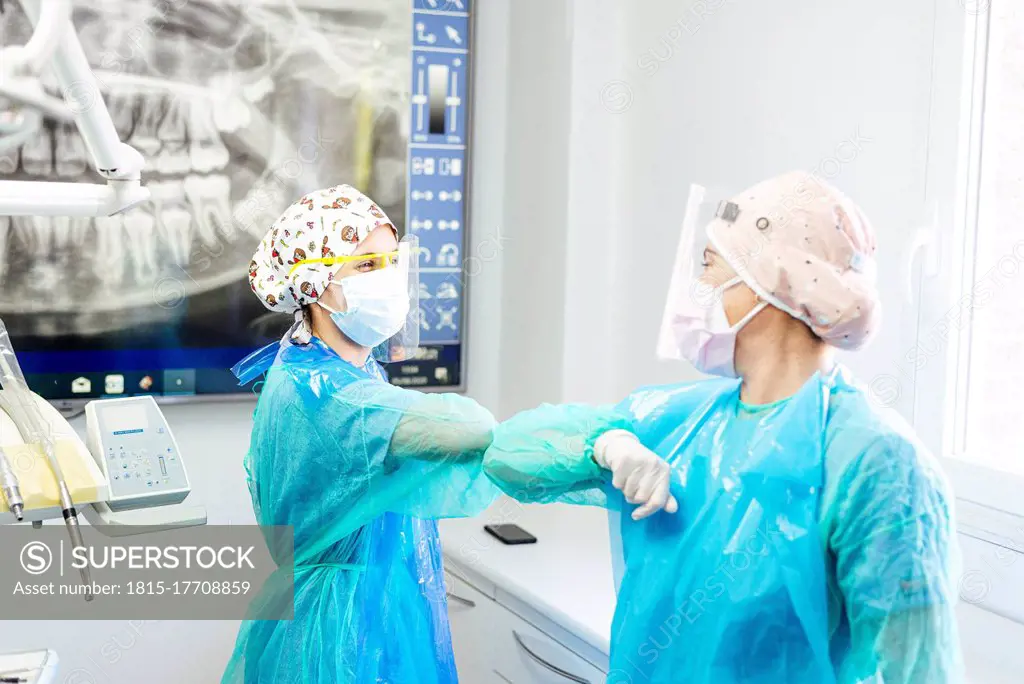 Dentist and assistant in protective workwear greeting with elbow at office