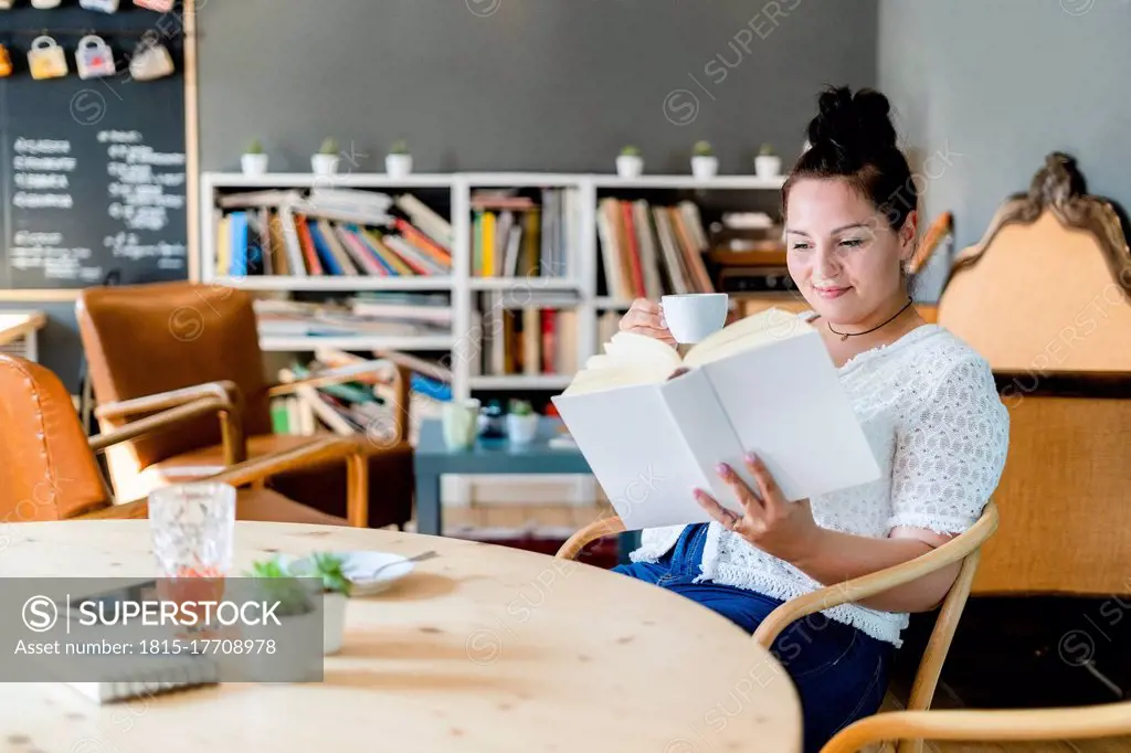 Young woman holding coffee cup reading book while sitting at table in cafe