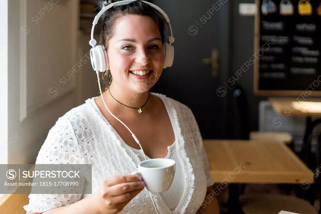 Close-up of smiling young woman holding coffee cup listening music through headphones in cafe