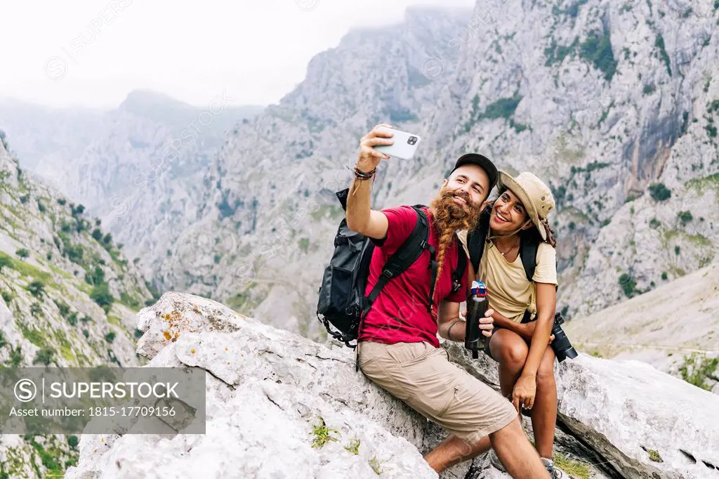 Smiling couple taking selfie while sitting on rock at Ruta Del Cares, Asturias, Spain