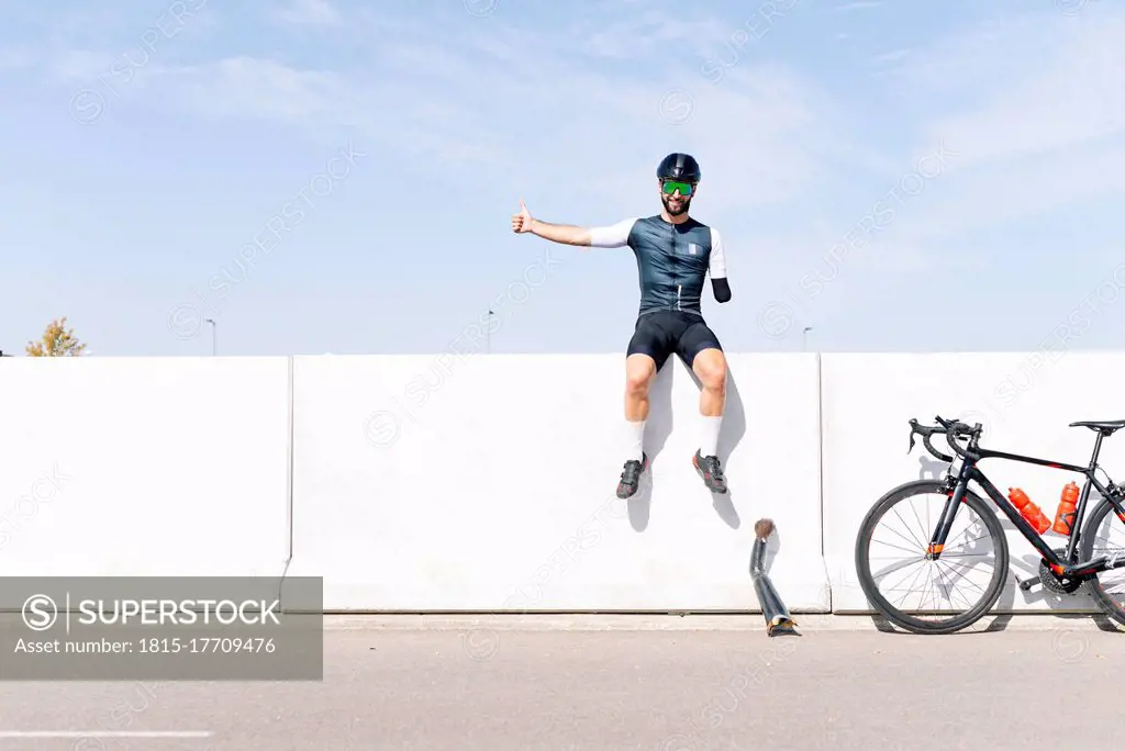 Male amputee athlete sitting on retaining wall against sky during sunny day