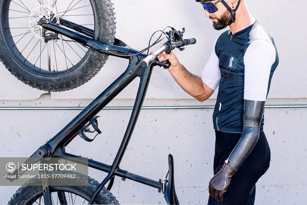 Male amputee athlete holding bicycle while standing by wall