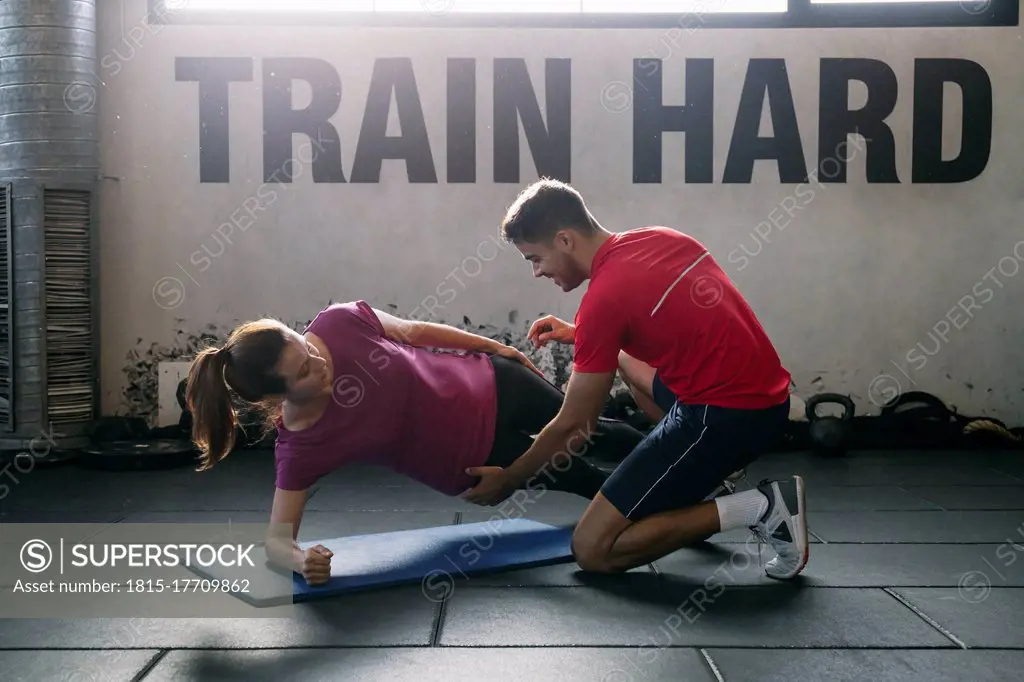 Fitness instructor assisting female athlete in practicing plank position on mat at gym