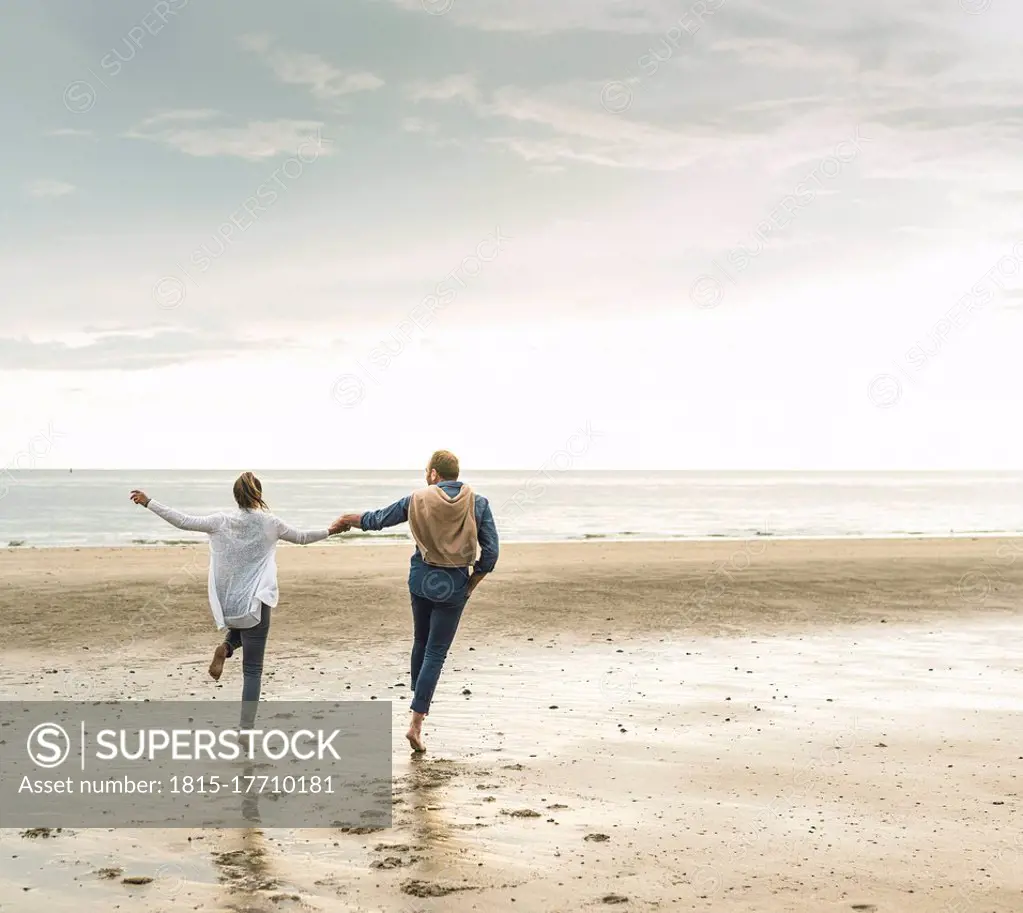 Cheerful mature couple holding hands while jumping at beach against cloudy sky