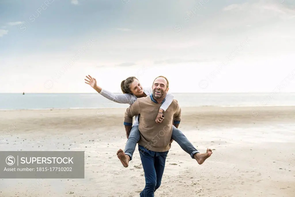 Happy mature man piggybacking woman while walking at beach against cloudy sky