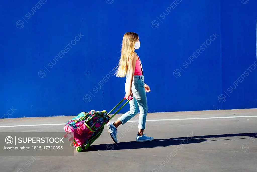 Blond girl walking with schoolbag on street against blue wall