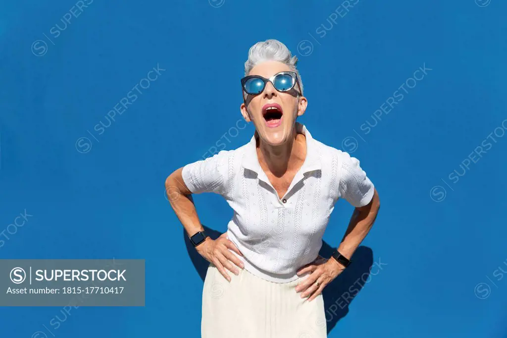 Woman with mouth open and hand on hip standing against blue wall during sunny day