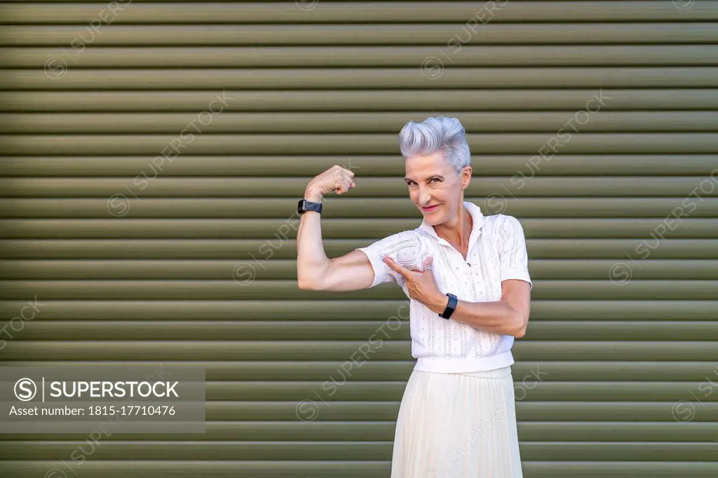 Active senior woman pointing at bicep while flexing muscles while standing against shutter