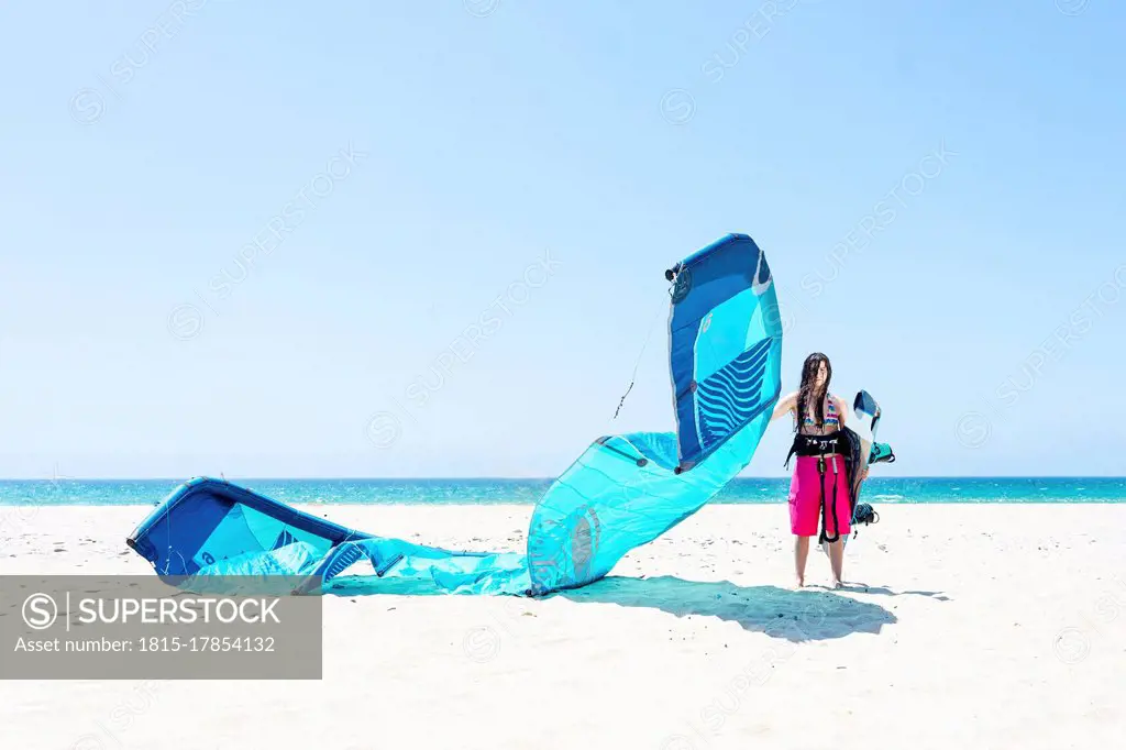 Young woman with kite and kiteboard standing at beach against clear sky during sunny day