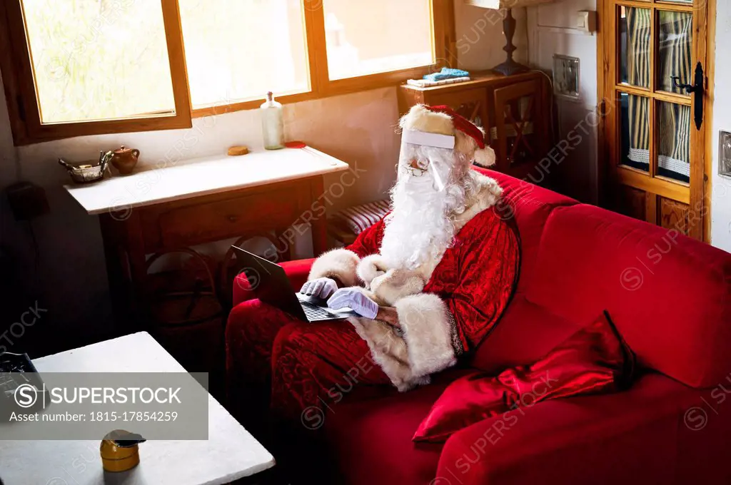 Man wearing Santa Claus costume using laptop while sitting on sofa at home