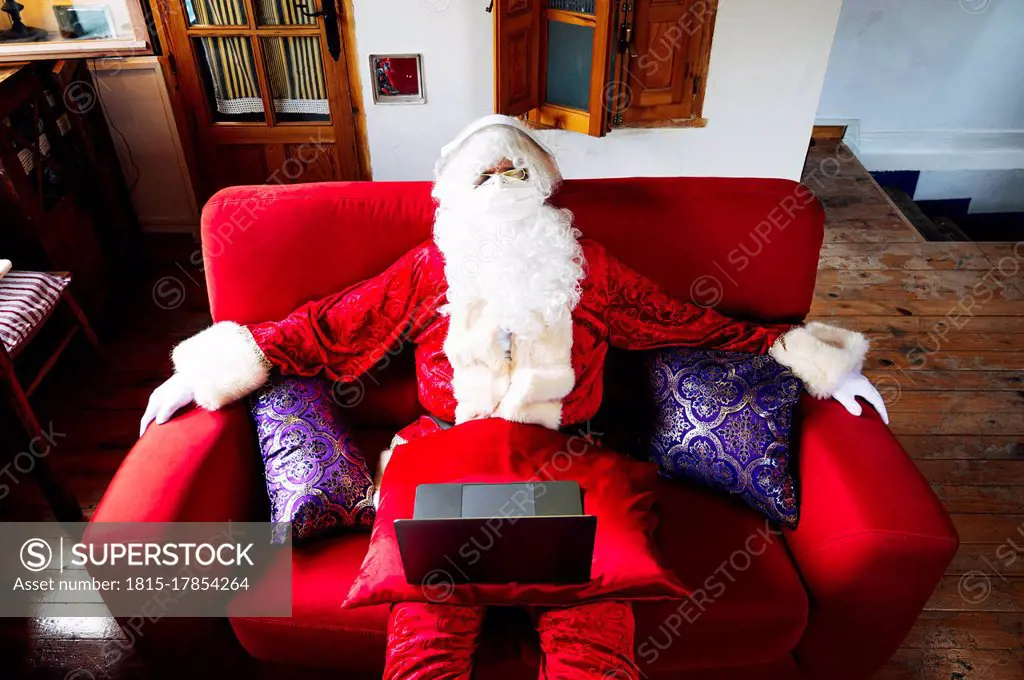 Man wearing Santa Claus costume with laptop relaxing on sofa at home