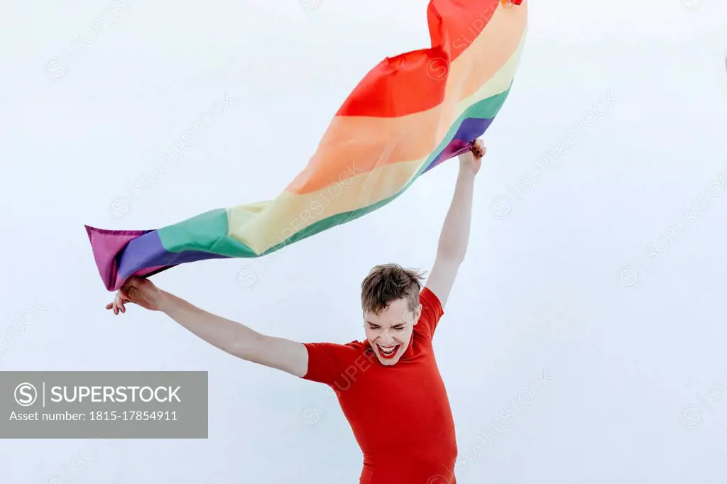 Non-binary person waving multi colored flag while standing against white wall