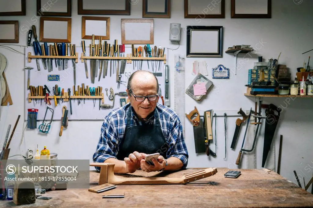 Man using mobile phone while leaning on workbench at workshop