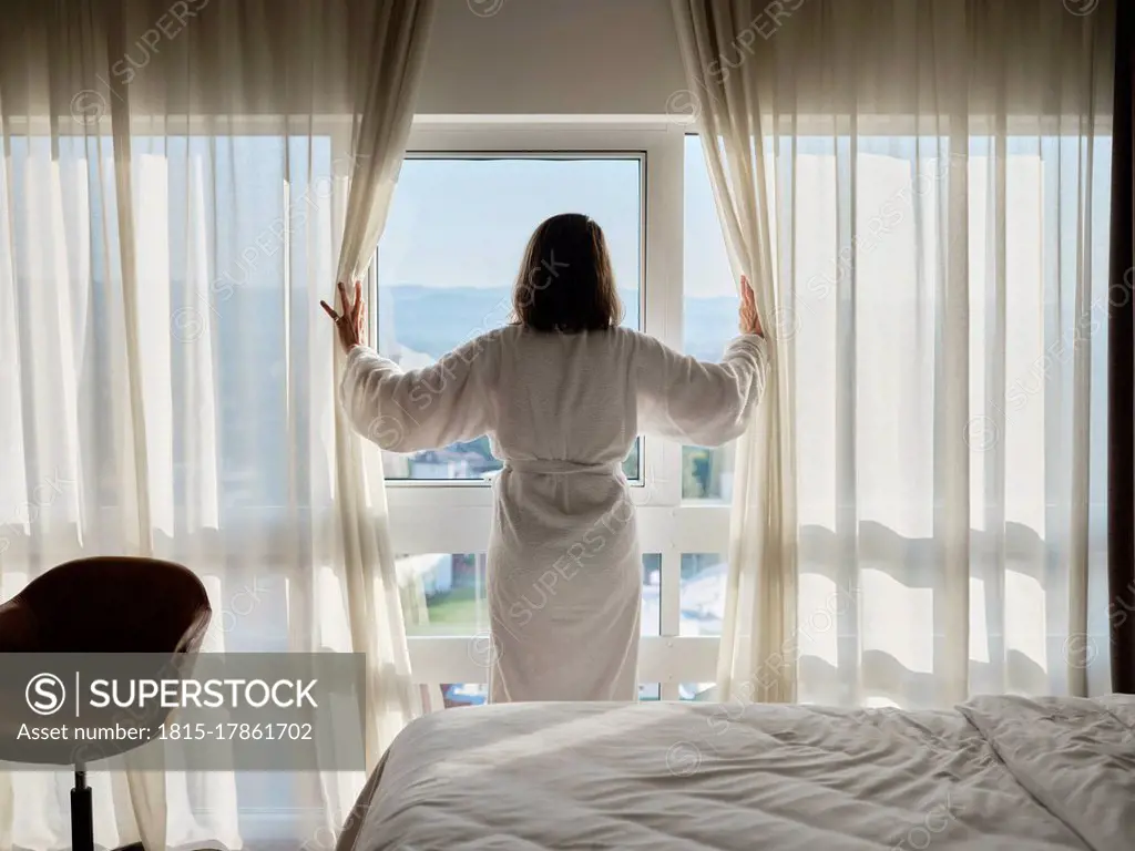 Woman opening white curtain while looking through window at luxury hotel room