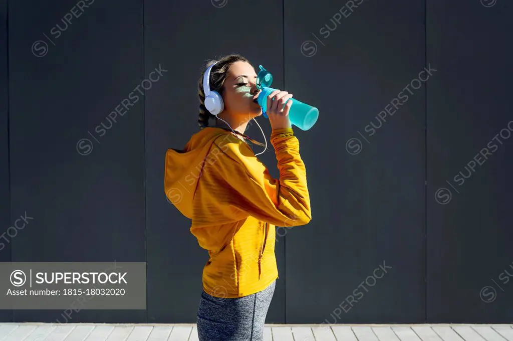 Female athlete wearing headphones drinking water while standing against gray wall