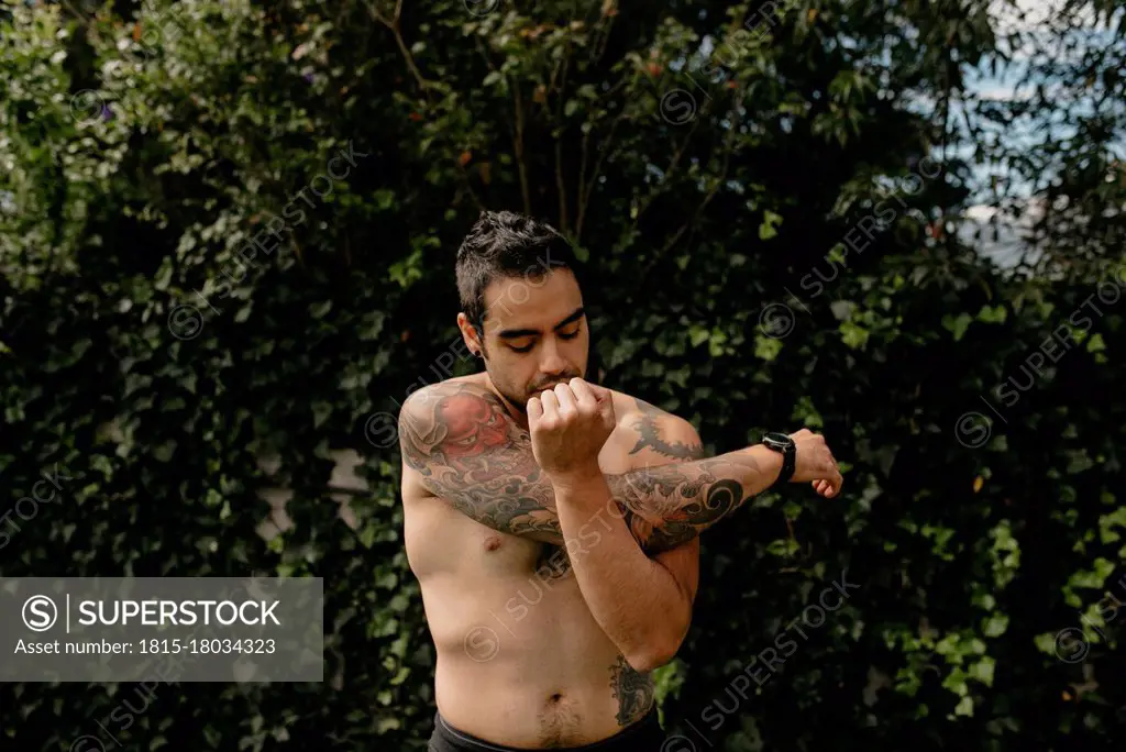 Shirtless handsome man with tattoo stretching arm while doing warm up exercise against plants at back yard