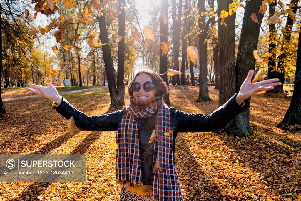 Smiling beautiful woman with arms outstretched throwing dry leaves while standing at park during autumn