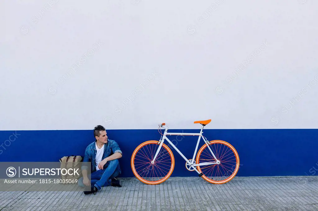 Man in casuals sitting on footpath by bicycle against wall