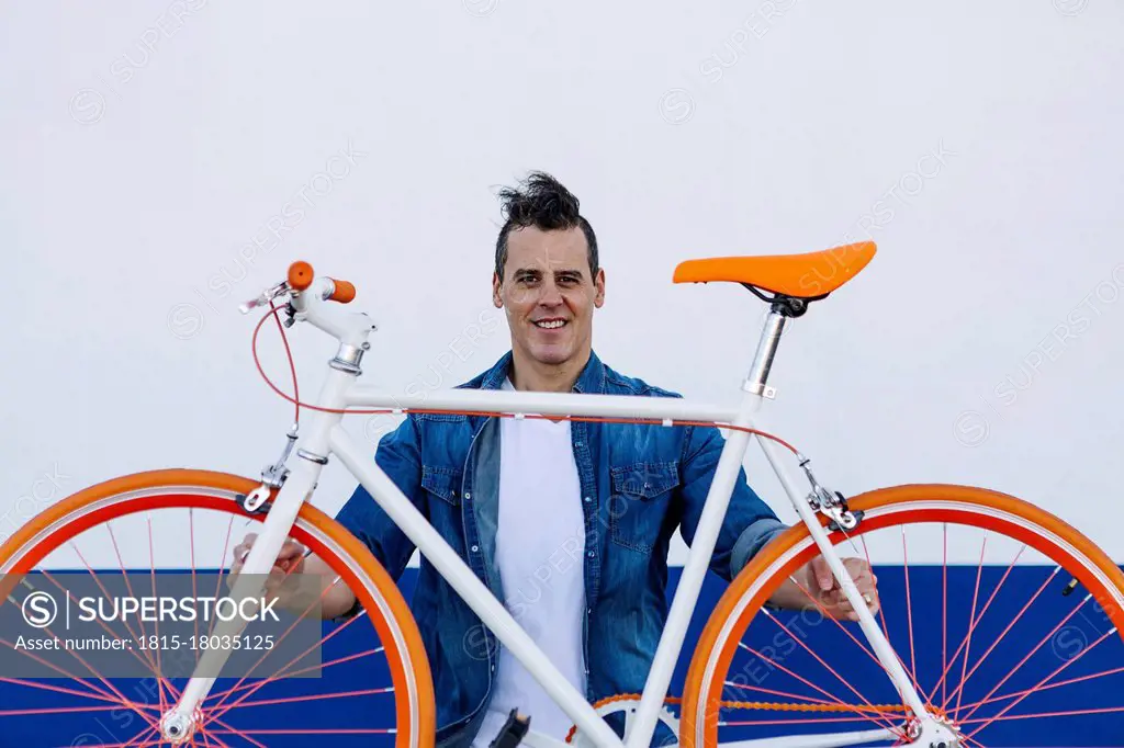 Smiling handsome man carrying orange bicycle against white wall