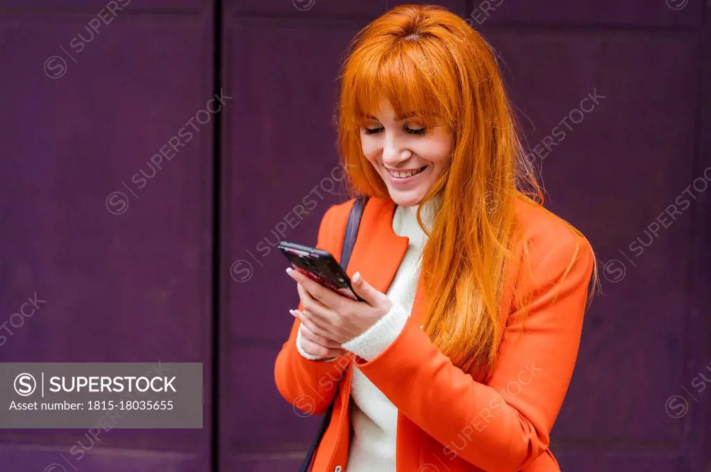 Smiling woman in jacket using mobile phone against wall