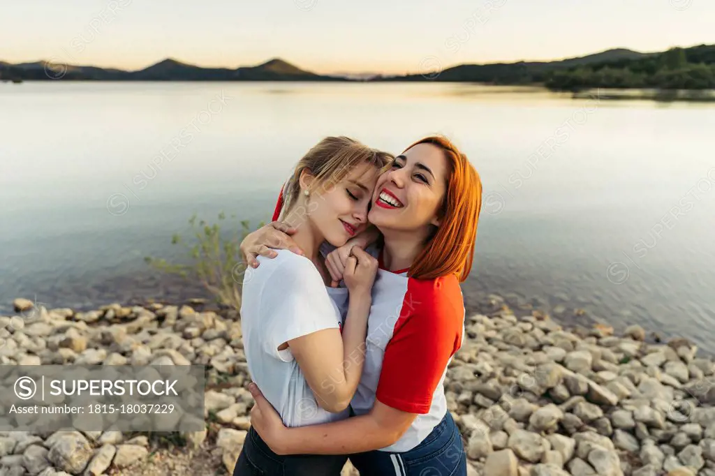 Happy woman embracing girlfriend while standing by lake during sunset