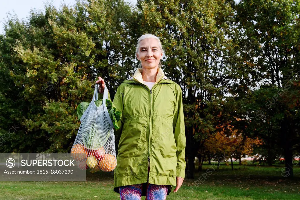 Mature woman holding mesh bag filled with fruits and vegetables at public park