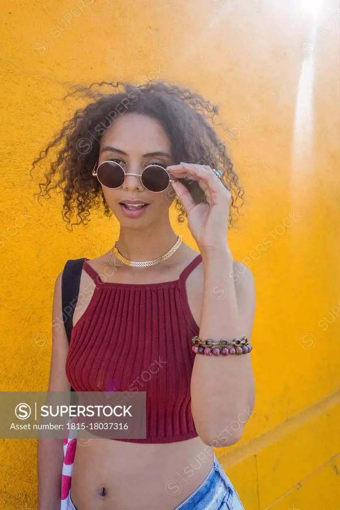 Fashionable young woman with curly hair holding sunglasses standing by yellow wall