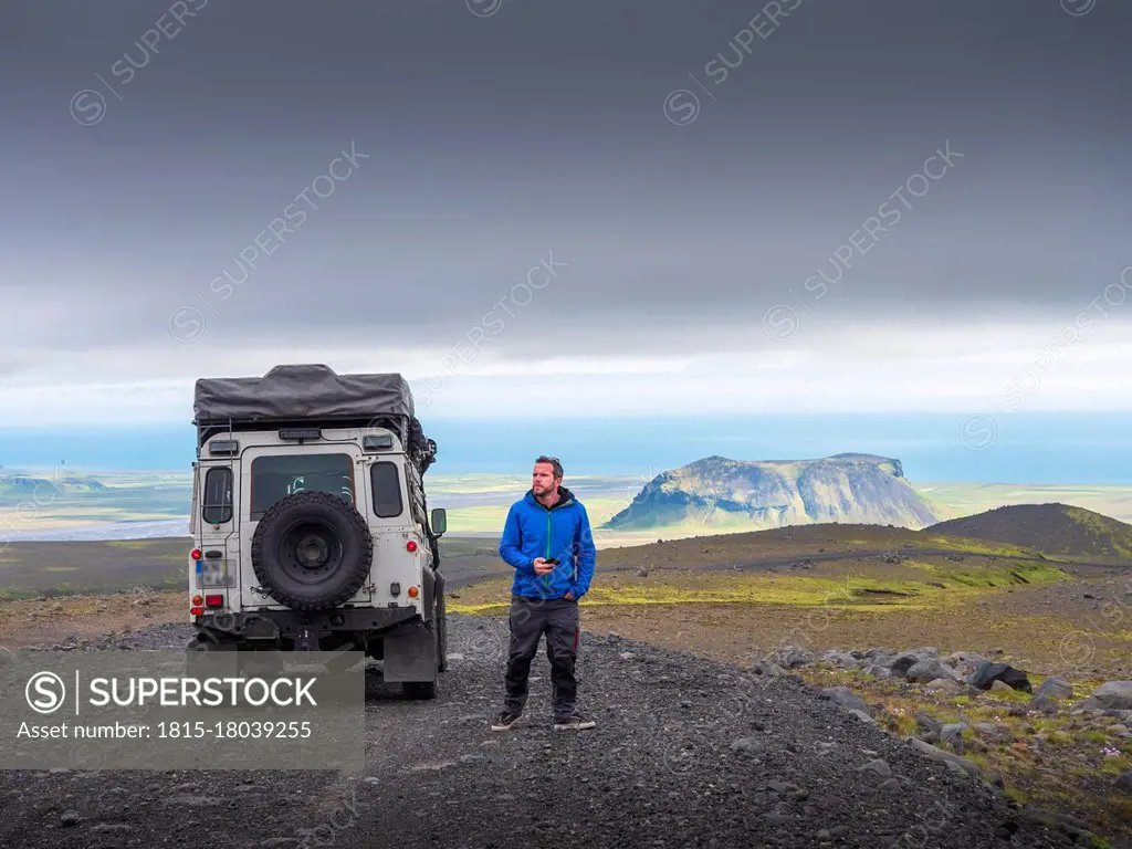 Male tourist looking away while standing by off-road vehicle on road against cloudy sky