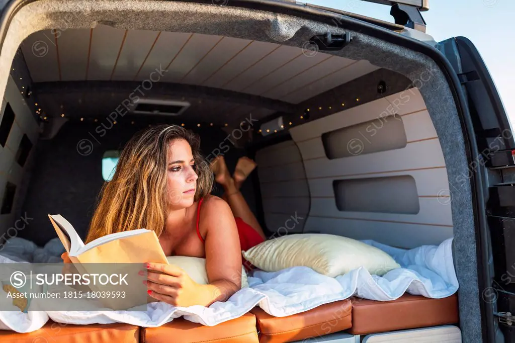 Woman with book looking away while lying in camper van at beach