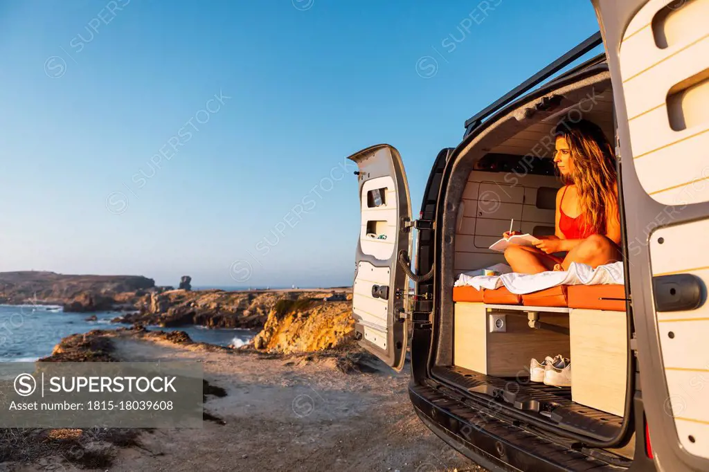 Young woman writing in book while sitting in camper van against at beach