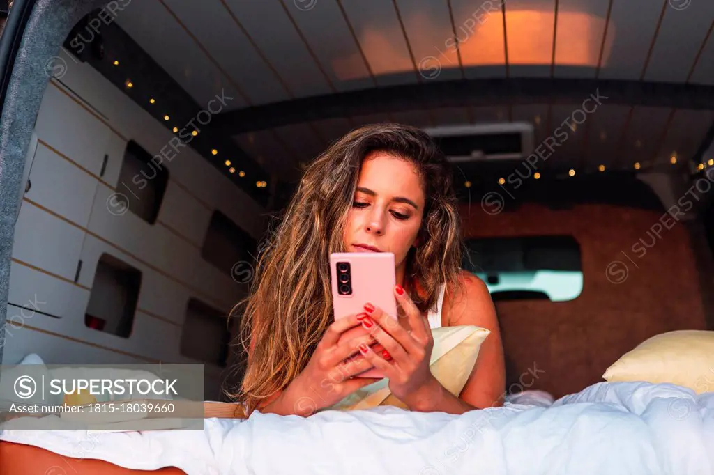 Beautiful woman using mobile phone while lying in camper van during sunset at beach